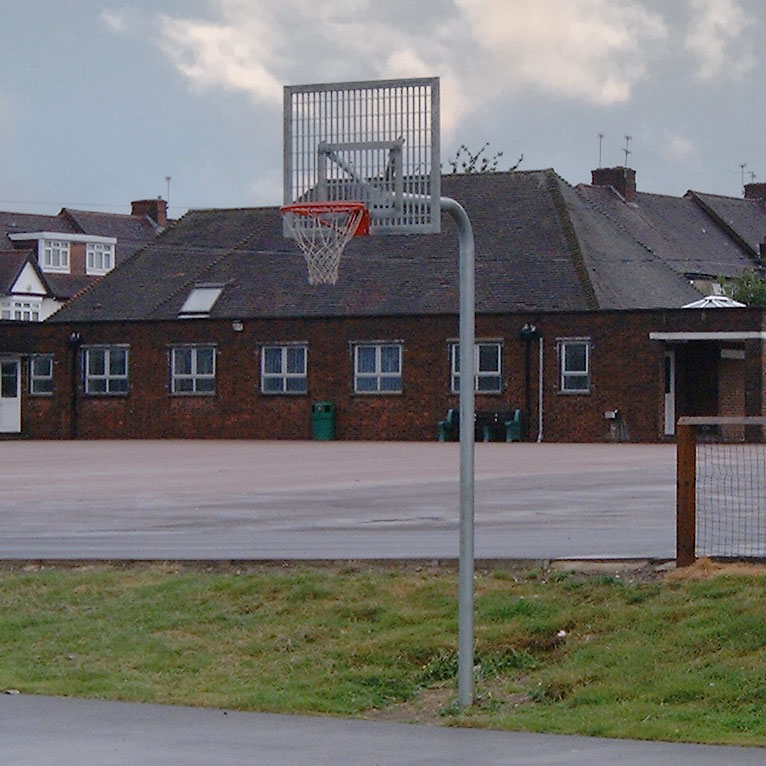 Outdoor school basketball hoop with a red rim and steel backboard, located on a tarmac court in front of a red-brick school building with pitched roofs and small windows, under a partly cloudy sky.