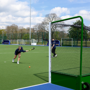 A green field hockey goal stands in an astroterf pitch, a young woman takes a practice shot